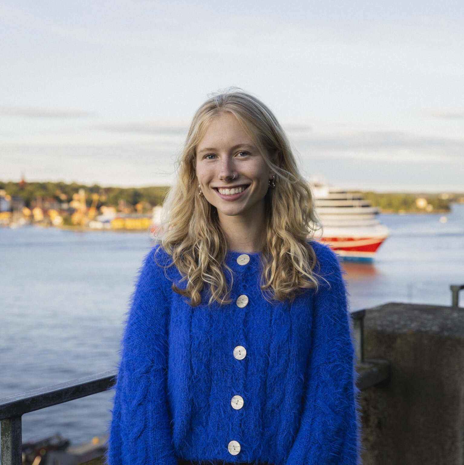 Headshot of Maggie Stevens on a balcony overlooking the Stockholm skyline, wearing a blue cardigan
