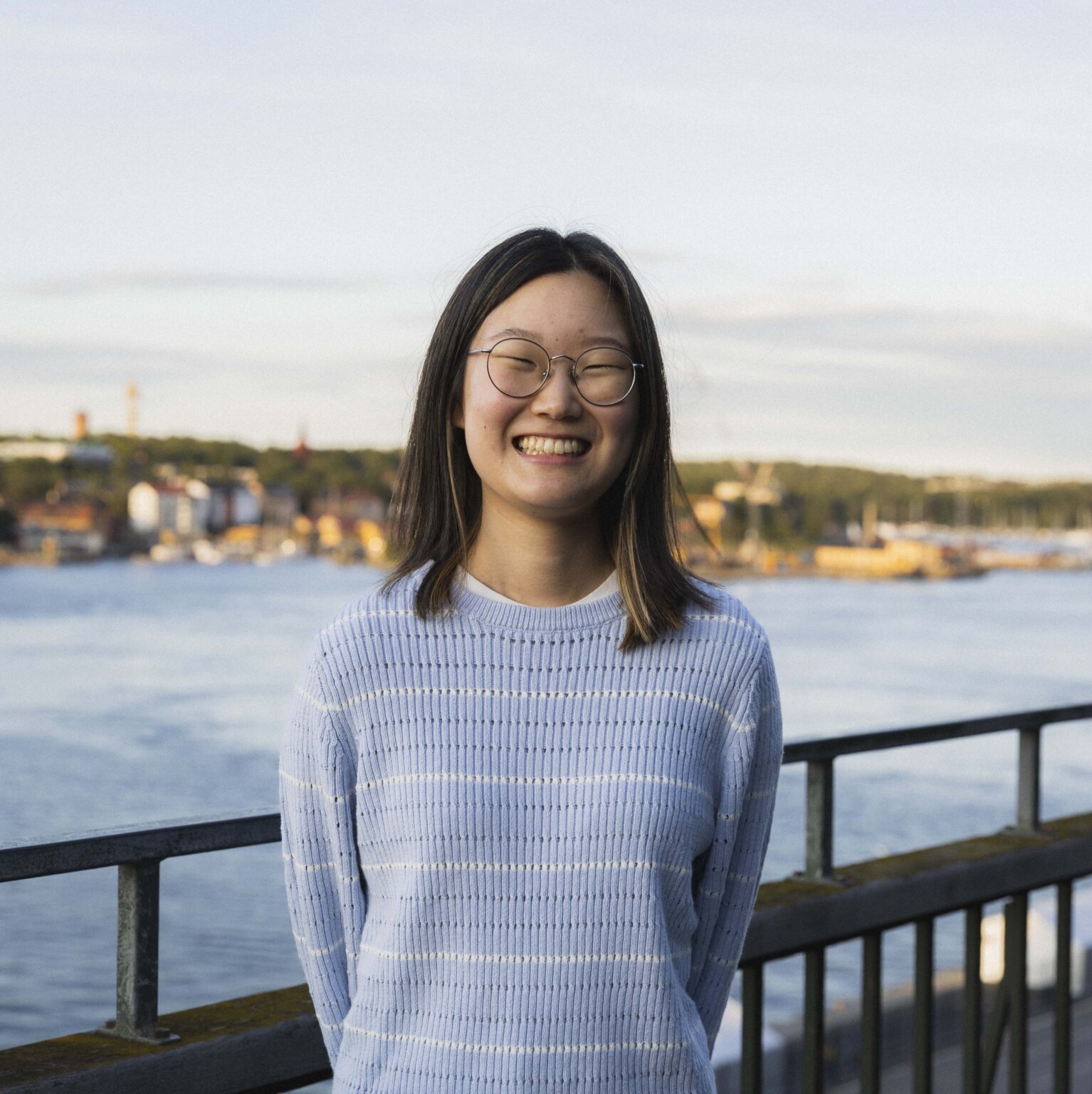 Headshot of Rayna Lee on a balcony overlooking the Stockholm skyline, wearing a light blue sweater