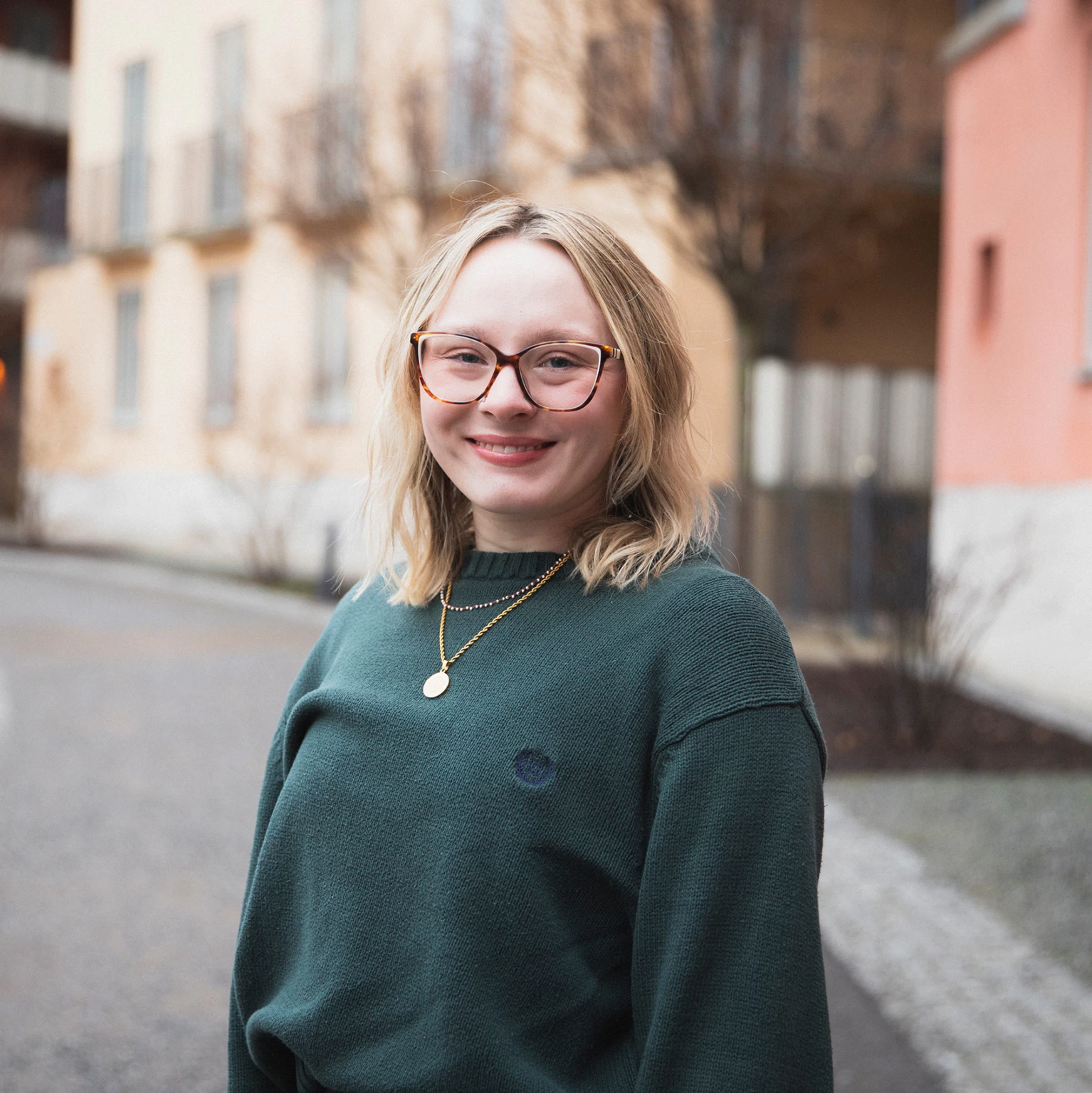 Headshot of Addison Ganey outdoors in front of yellow and orange buildings, wearing a dark green sweater
