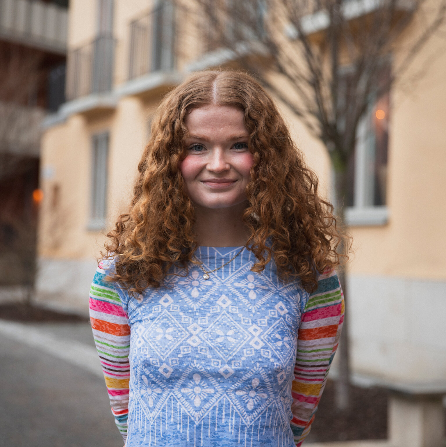 Headshot of Diane Meyer outdoors in front of a yellow building, wearing a colorful top