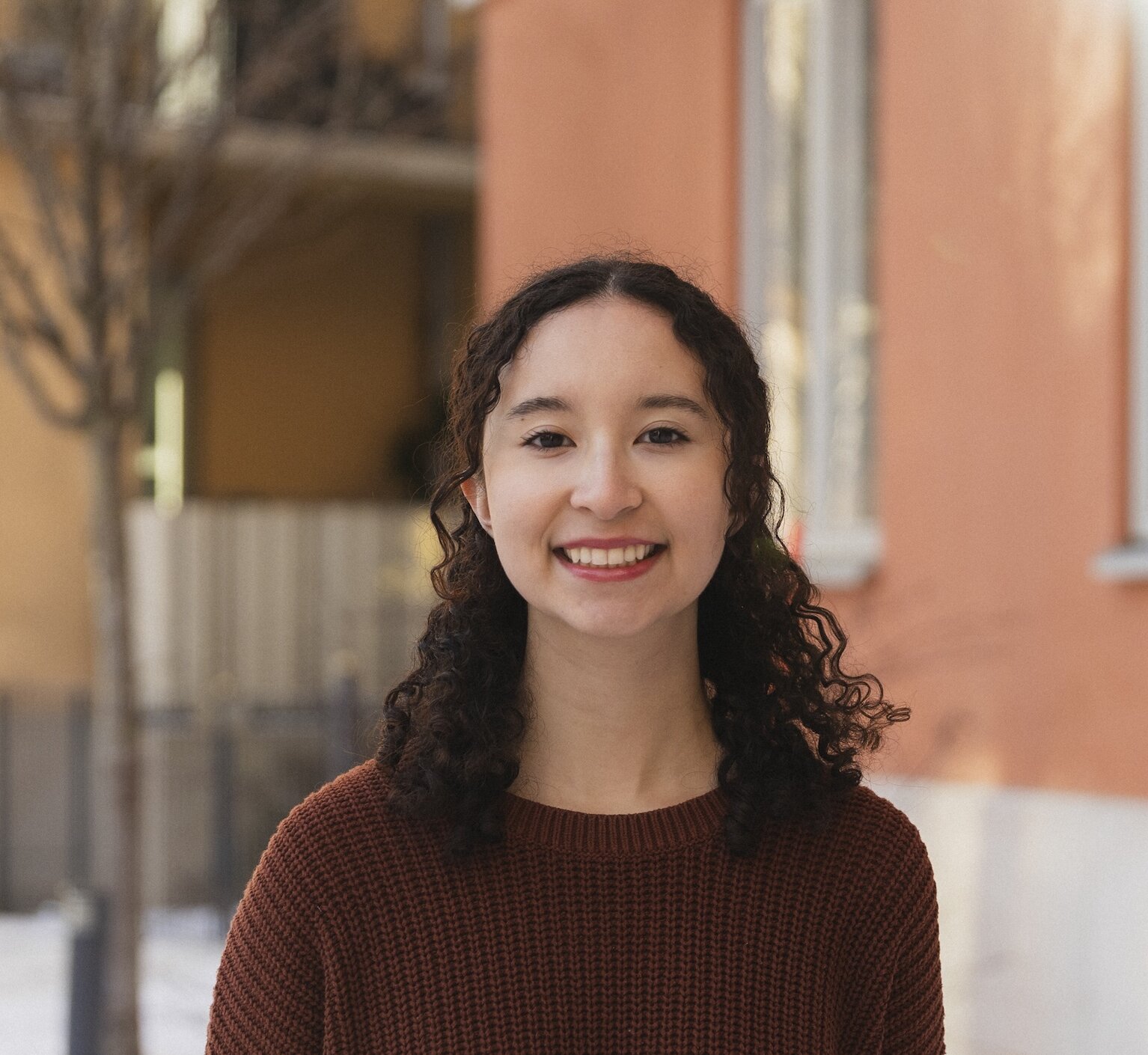 Headshot of Gaby Lopez outdoors in front of yellow and orange buildings, in a brown sweater.