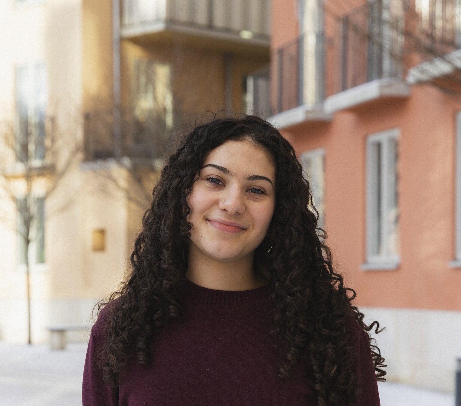 Headshot of Lizette Guajardo outdoors in front of yellow and orange buildings, in a burgundy sweater.
