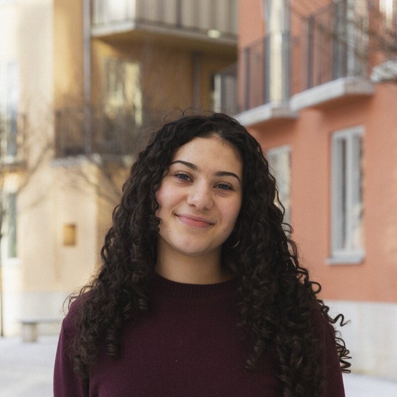 Headshot of Lizette Guajardo outdoors in front of yellow and orange buildings, in a burgundy sweater.