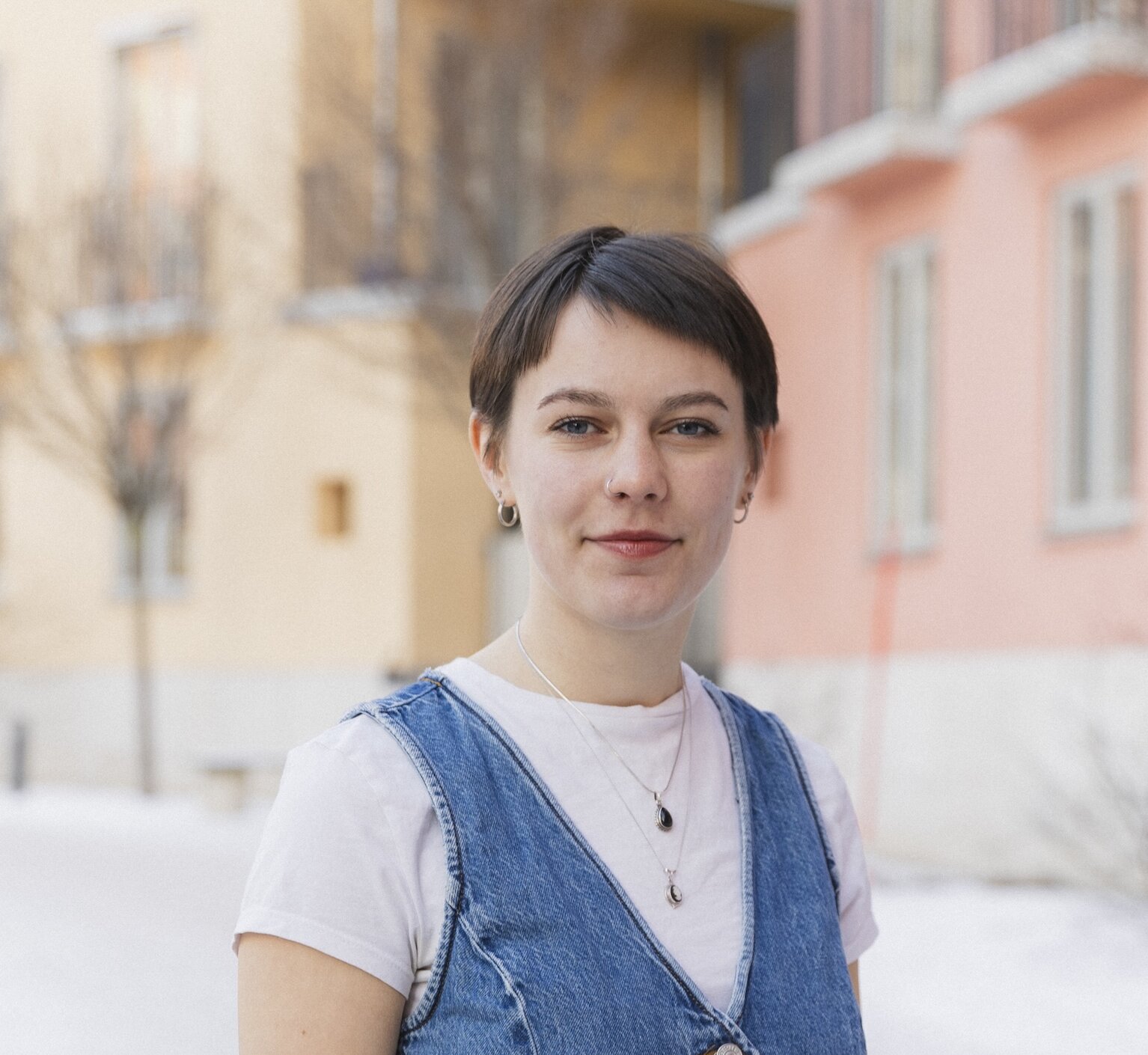 Headshot of Ruth Dikkers outdoors in front of yellow and pink buildings, in a white tshirt and denim vest.