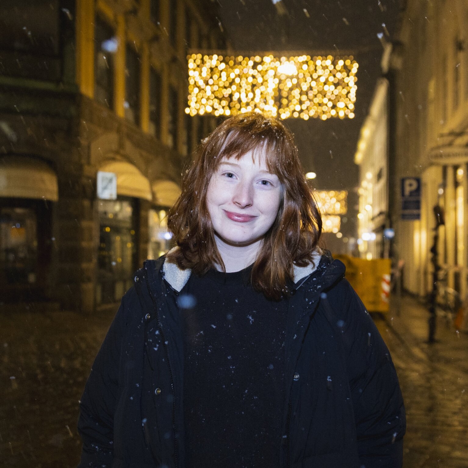 Night time headshot of Clara Steege on a Copenhagen street in the snow, wearing a black jacket
