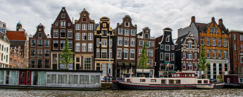 Photo of the canals with boats passing by with multiple apartments behind it. The apartments have the traditional Dutch four-second facades.