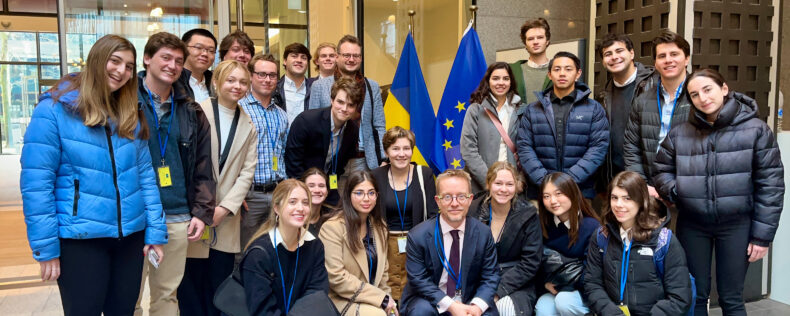 Students posing with their professor and a guest in front of the EU flag. They are a part of a European politics class.