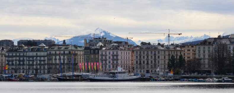 Overlooking Geneva with water in the foreground and historic buildings. With mountains covering the horizon.