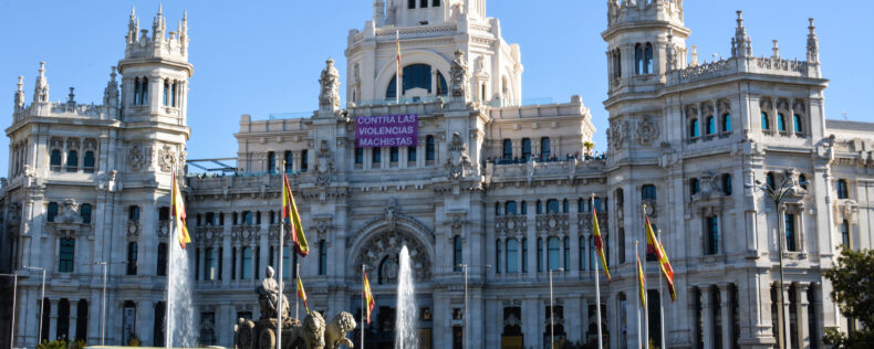 The front of a town hall building in Madrid. There is a historic statue surrounded by a foundation and Spainish flags.