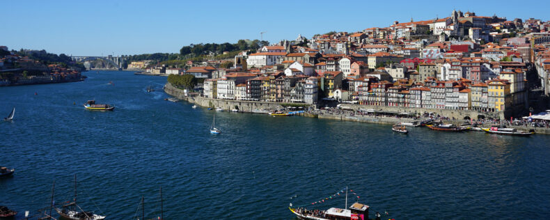 A view of Porto with The Dom Luís I Bridge in the background to the left. A boat cruising in the water in the foreground with the hills of the city behind it.