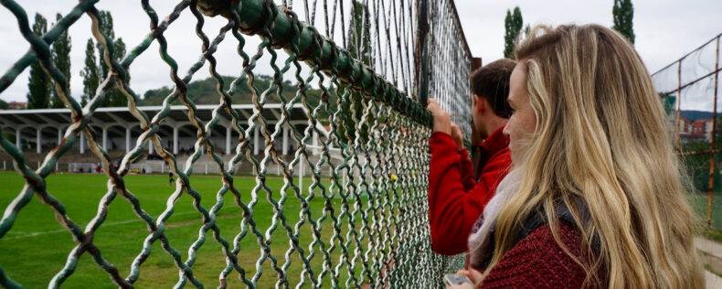 Two students looking through a green chain-link fence. The paint is peeling and the day is overcast. The students are both wearing red.