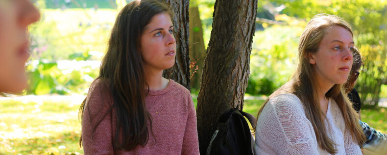 Two students attentively listening to a lecture while in the park. Lots of greenery in the background.