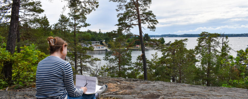 A student reflecting and taking notes on top of a rock over looking a Swedish lake.