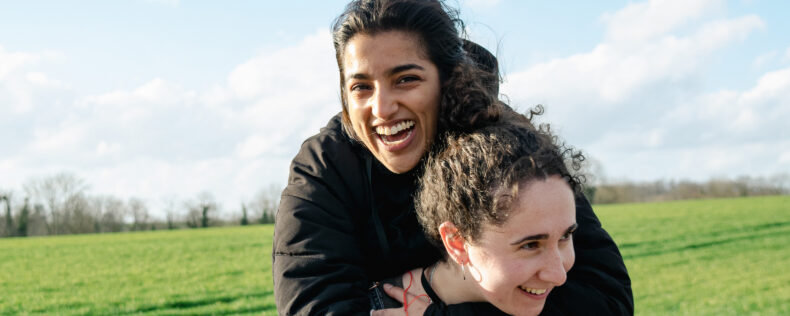 One student giving another student a piggy back ride in a Swedish field. Both are laughing and full of joy.